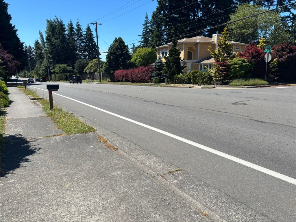 View of a residential street with a sidewalk, mailbox, and a house in the background, surrounded by trees and shrubs under a clear blue sky.